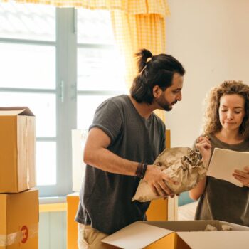 Free Cheerful young man and woman smiling while unpacking carton boxes with belongings in new apartment during relocation and looking at paper Stock Photo